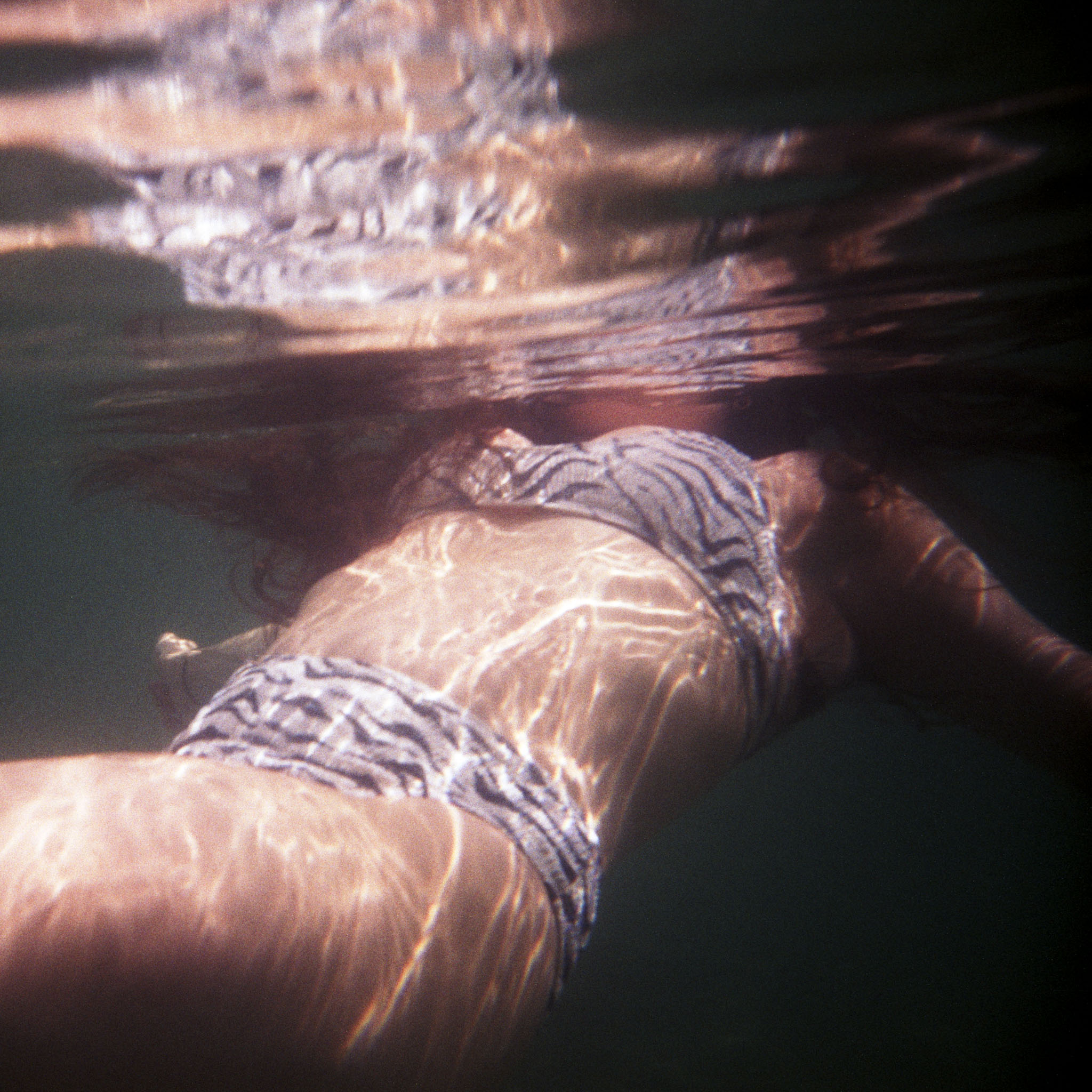 Swimmer at the waterline of an ocean pool seen from below, caustic light across patterned swimsuit — 35mm Kodak Portra 400